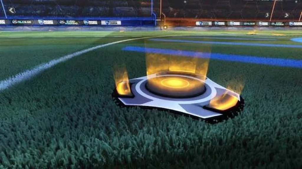 Boost no Rocket League: o que é, como funciona e por que ele é tão importante 1 Pad de boost pequeno do Rocket League sobre o gramado da arena, com energia dourada ativa e campo dividido entre os lados azul e laranja, destacando um ponto de recarga de boost em alta qualidade.