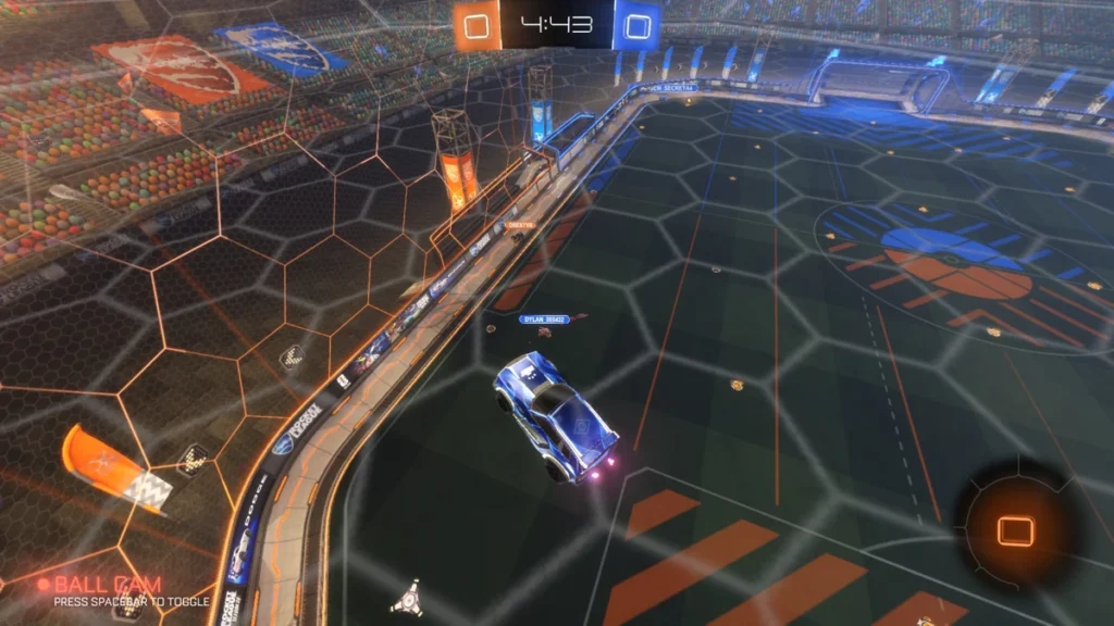 Carro azul em disputa aérea durante partida de Rocket League, próximo à parede lateral da arena, em lance competitivo com placar empatado em 0 a 0 e foco na movimentação tática.
